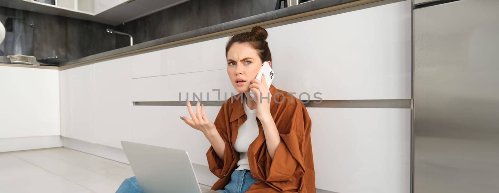 Woman with frustrated and confused face talking on mobile phone, shrugging and frowning, hear bad news over the telephone, sits on floor with laptop, calls customer support by Benzoix