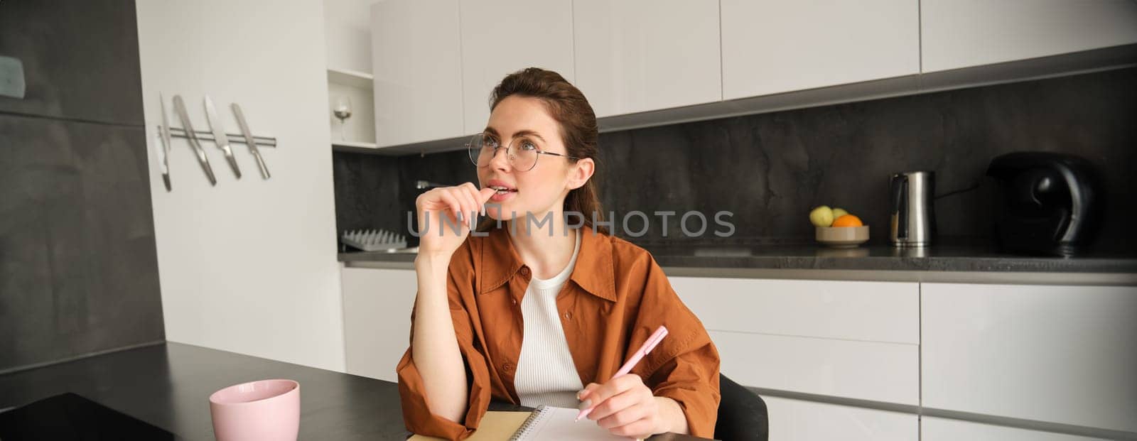 Young creative woman, writer making notes, holding pen, looking aside with thinking face, sitting in kitchen at home, doing homework.
