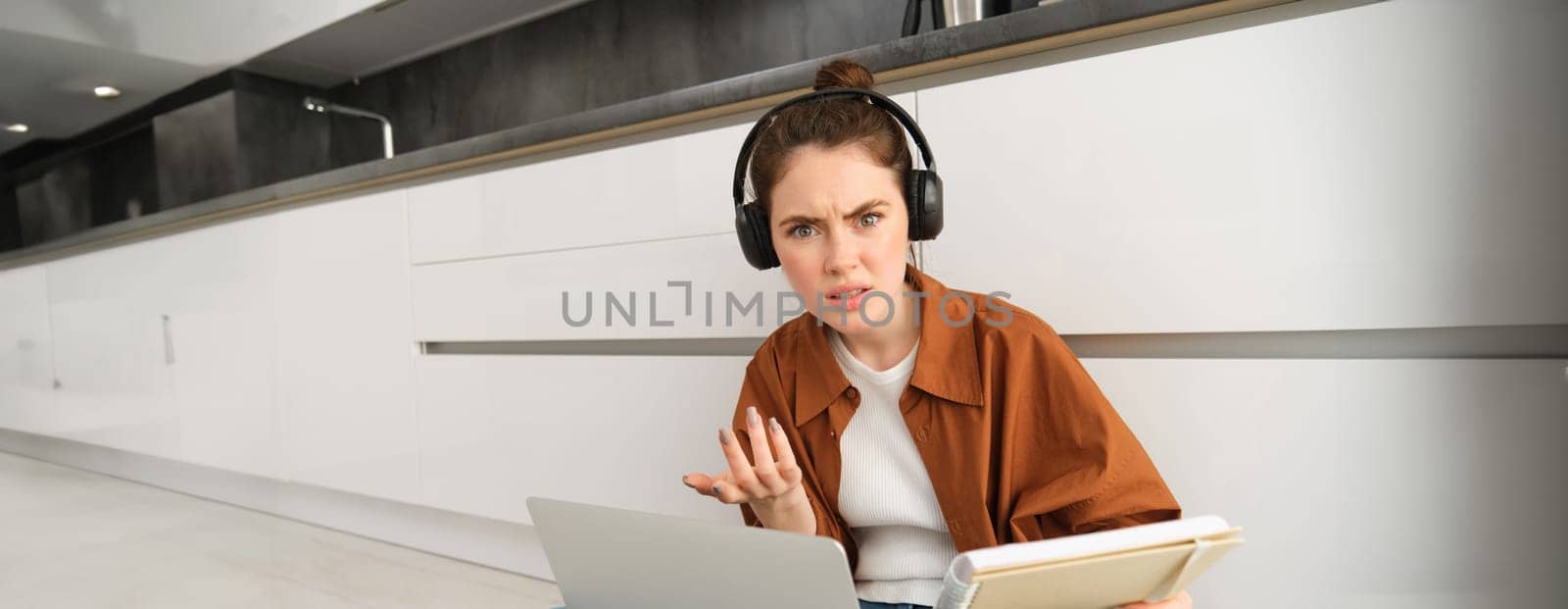 Confused and frustrated woman in headphones, student doesnt understand something while doing homework on laptop, looking annoyed at camera by Benzoix