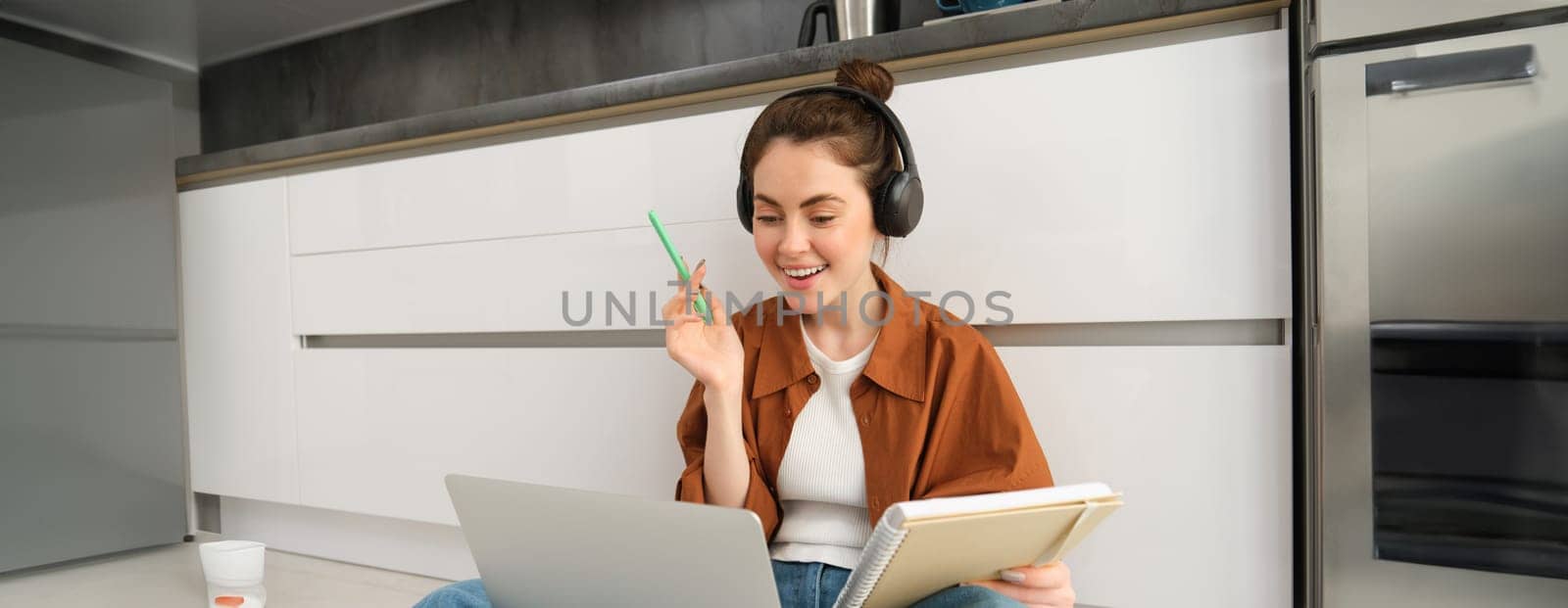 Woman studies, does homework at home, connects to online lesson, sits on floor with laptop, writes in notebook, works in headphones by Benzoix