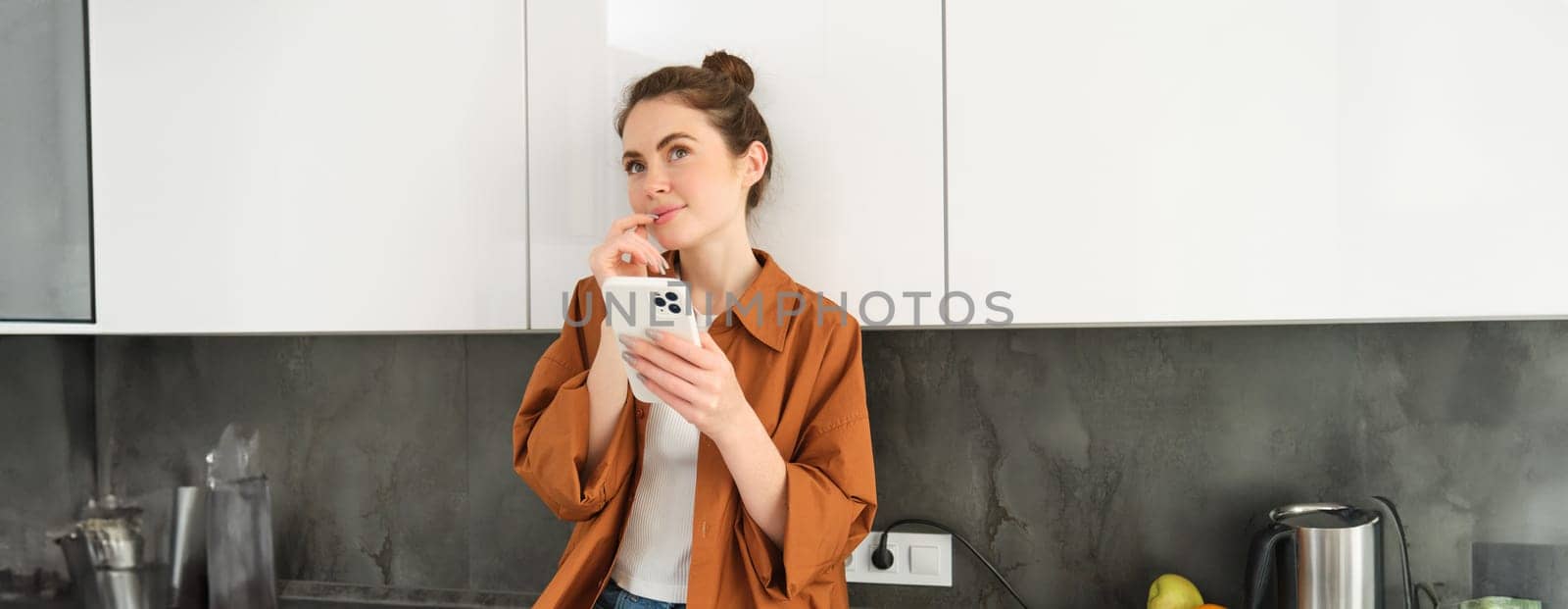 Portrait of woman online shopping using smartphone app, sitting in kitchen with thinking face, holding mobile phone by Benzoix