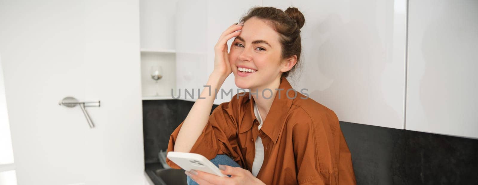 Portrait of young woman with mobile phone, browsing social media, online shopping, sitting on kitchen counter, smiling and looking happy.