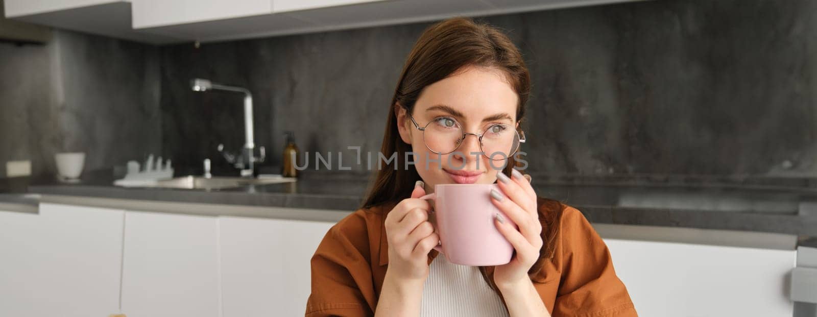 Portrait of beautiful young woman in glasses, drinking coffee, holding cup and enjoying her tea in kitchen by Benzoix