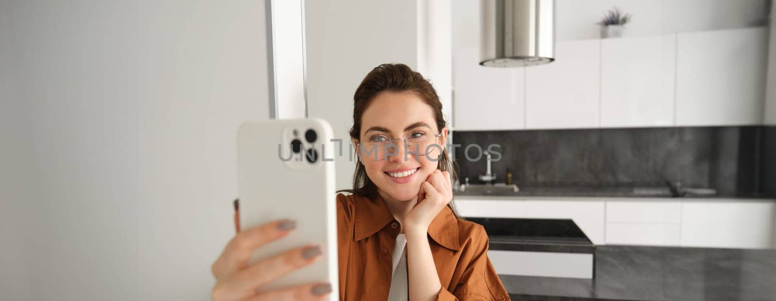 Beautiful young woman in glasses taking selfie in her flat, using smartphone, posing for photo, standing with mobile phone and smiling by Benzoix