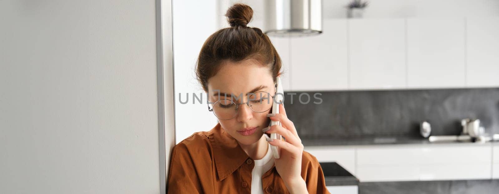 Portrait of young sad woman in glasses, talking on mobile phone, listening to voice message on smartphone with upset, concerned face expression, standing at home, leaning on wall.