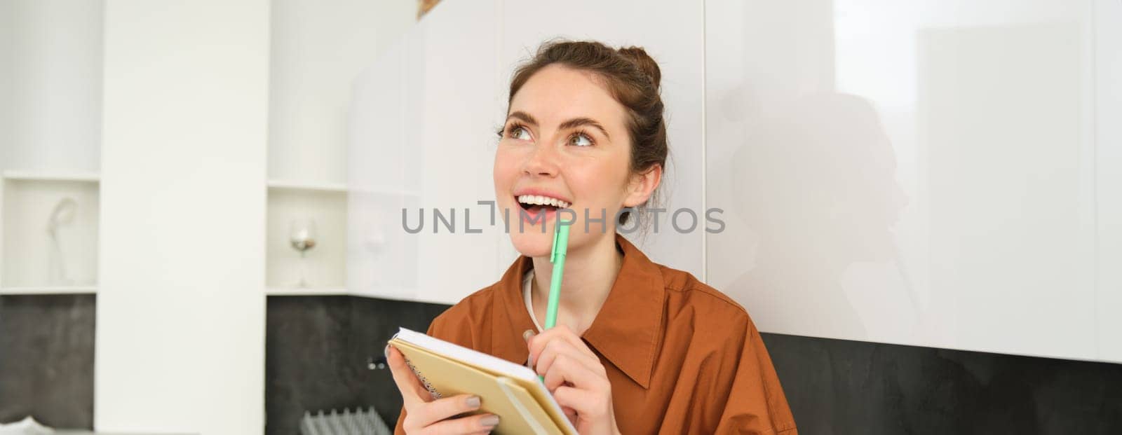 Portrait of young smiling woman with creative thoughts, looking up with inspired face, holding pen and notebook, writing down her ideas.