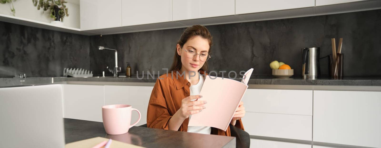 Portrait of woman working from home, sitting in kitchen with documents, opens folder, drinks tea by Benzoix