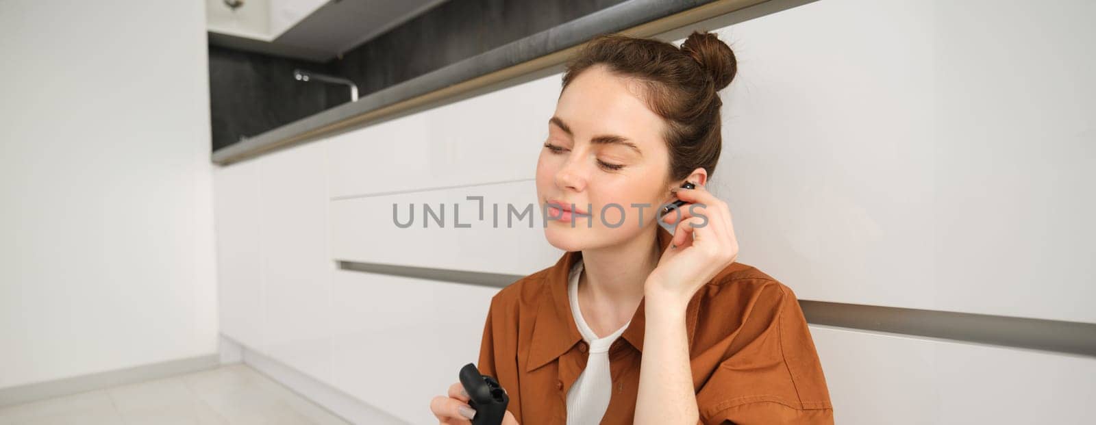 Portrait of young smiling woman listens to music in her black wireless earphones, using headphones to enjoy sound quality of song, sits on kitchen floor.