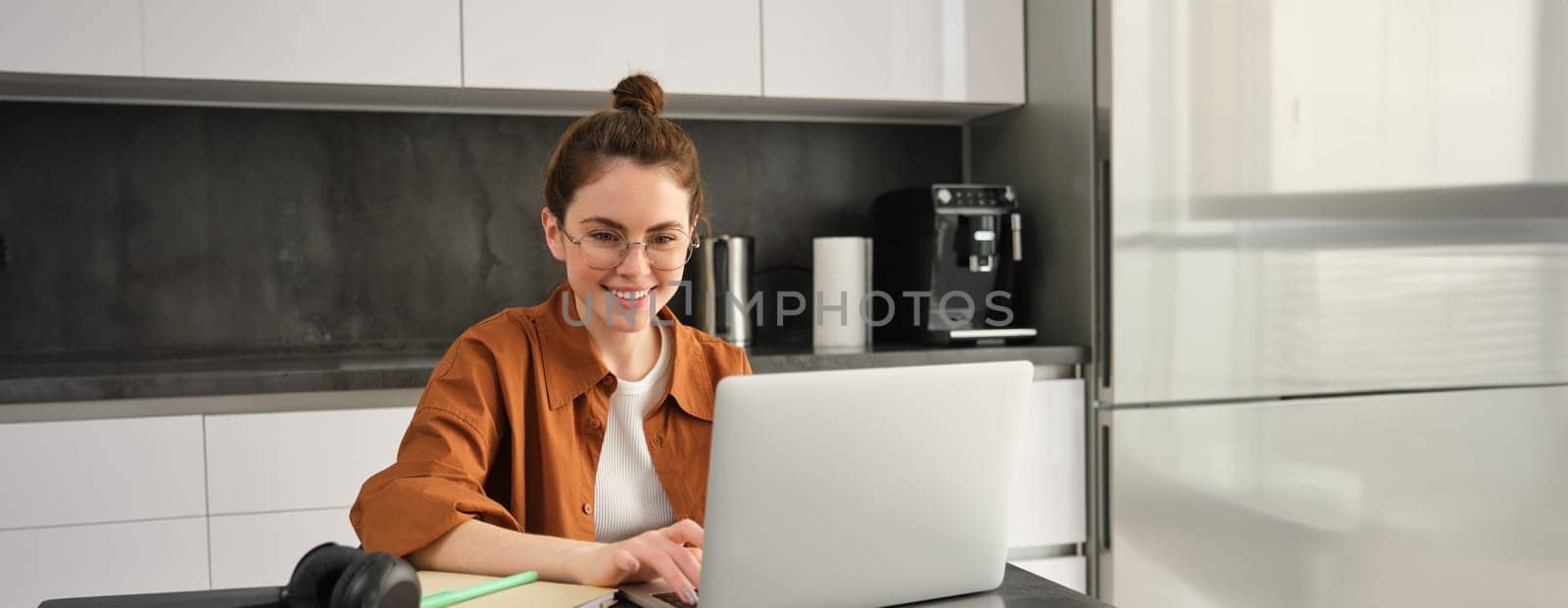 Portrait of young woman working from home, sets up workplace in kitchen, using laptop. Student doing homework on computer.