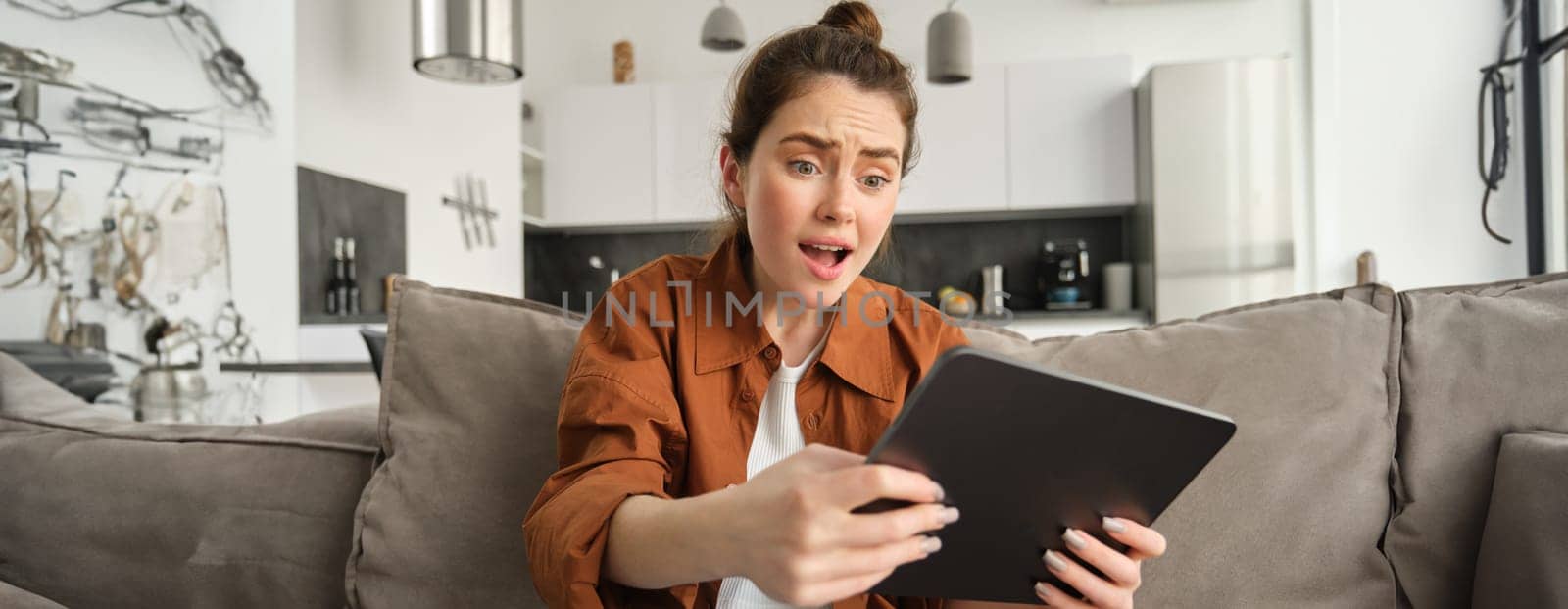 Portrait of woman with worried face looking at her digital tablet, losing in video game, sitting on couch at home. Technology and leisure concept