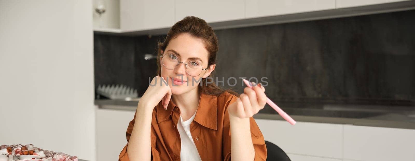 Close up portrait of smiling beautiful woman writing with pen, sitting in kitchen and doing homework, student studying, sitting in kitchen, wearing glasses.