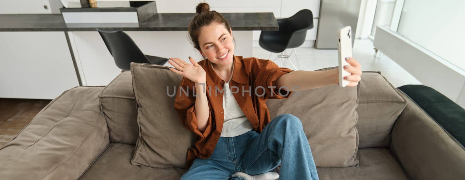 Photo of smiling young woman sitting on couch, posing on sofa in living room, taking selfies for social media.