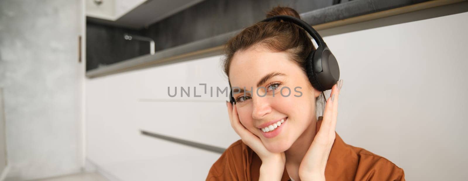 Close up portrait of beautiful brunette woman, listens to music in wireless headphones, has new earphones on her head, smiling and looking happy by Benzoix