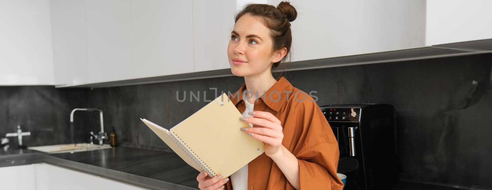 Portrait of gorgeous young woman reading, student standing in the kitchen with planner, doing homework, revising for exam.