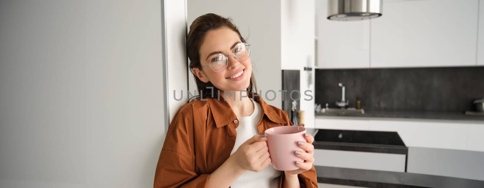 Portrait of cosy, beautiful and relaxed young woman at home, standing and leaning on wall with cup of coffee, drinking tea with thoughtful face expression by Benzoix