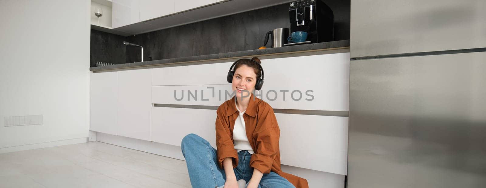 Cute brunette woman on kitchen floor, wears headphones, listens music and smiles, enjoys sound quality.