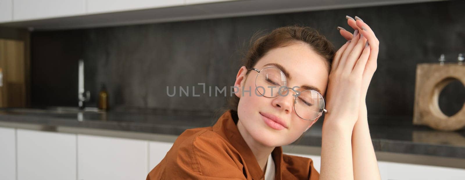 Portrait of beautiful, tender young woman in glasses, close her eyes and smile, thinking of something, dreaming, sitting in the kitchen by Benzoix