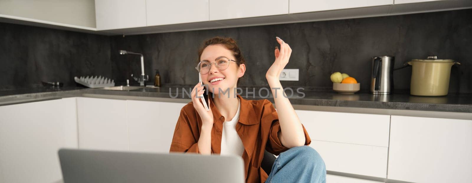 Image of beautiful modern woman working from home, studying in kitchen with laptop, talking on smartphone, calling someone on telephone.