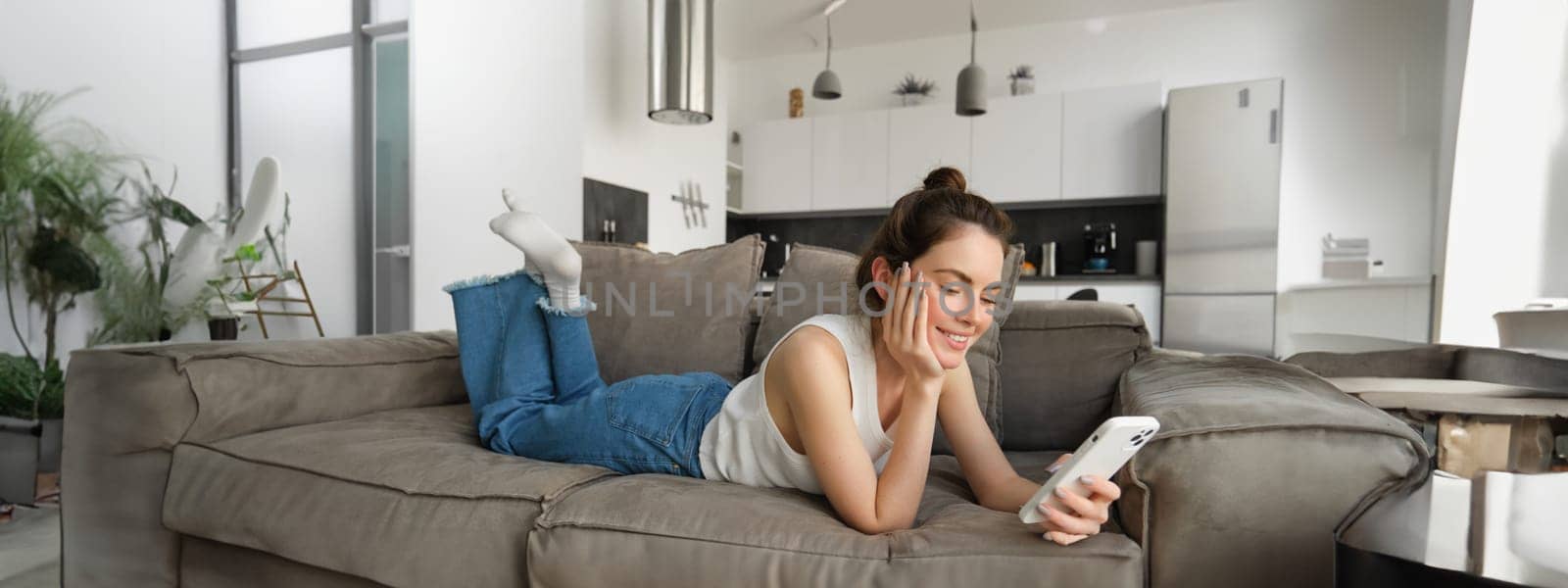 Portrait of cute young woman with smartphone, lying on couch and resting at home, scrolling social media feed, online shopping, reading something on mobile phone.