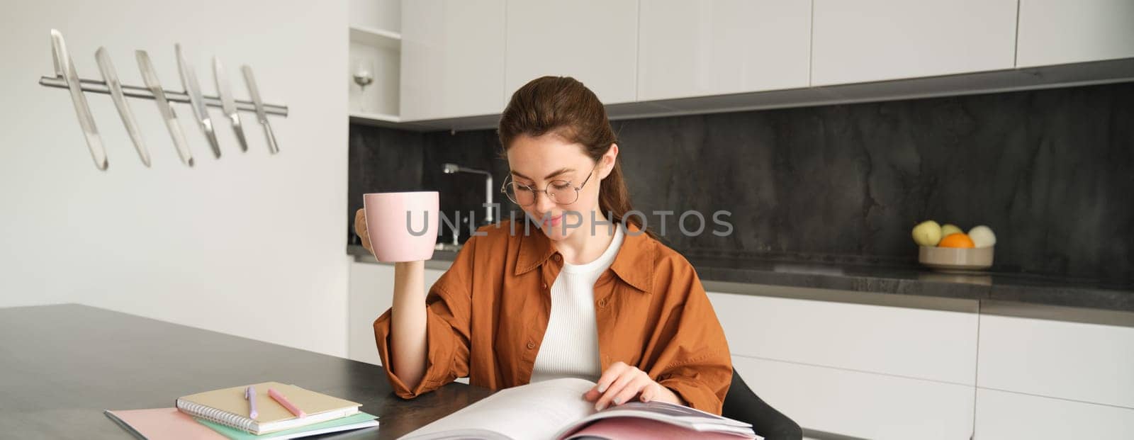 Portrait of beautiful student, woman in glasses reading and drinking coffee at home, studying and revising for lesson, doing homework by Benzoix