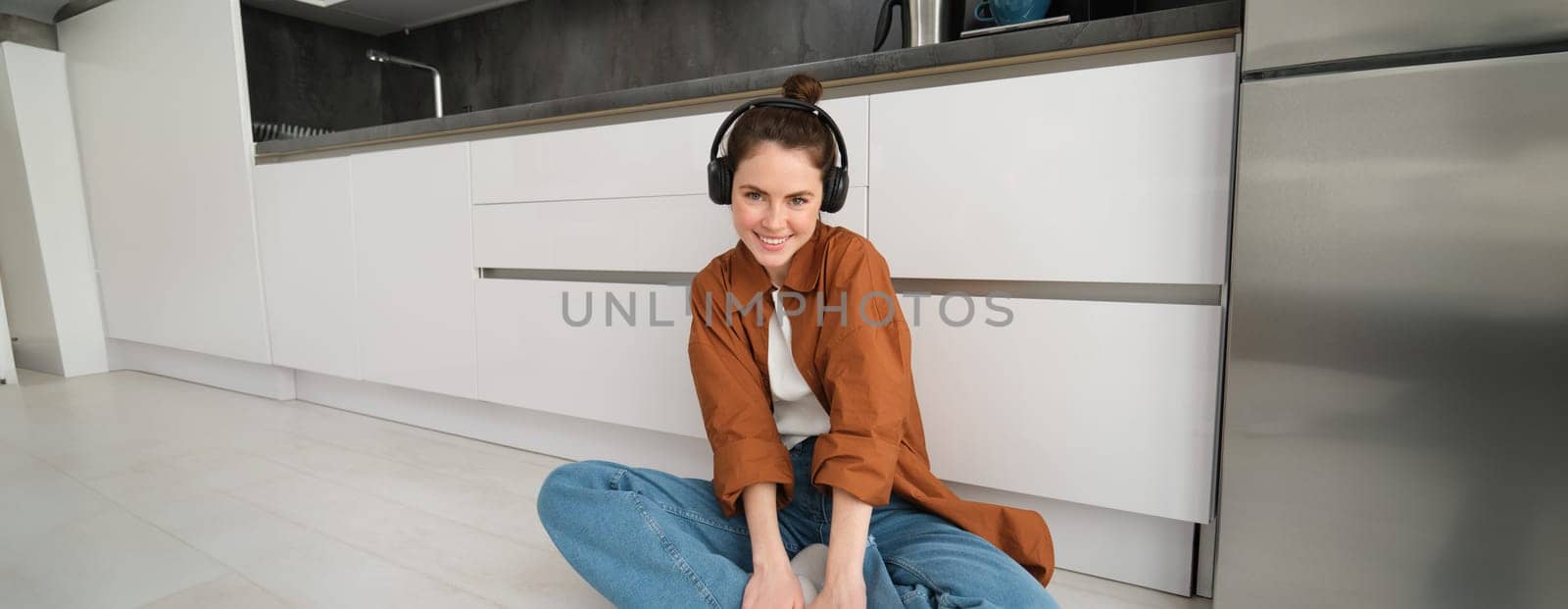 Portrait of young beautiful woman in wireless headphones, sitting on kitchen floor, vibing with favourite music in earphones.