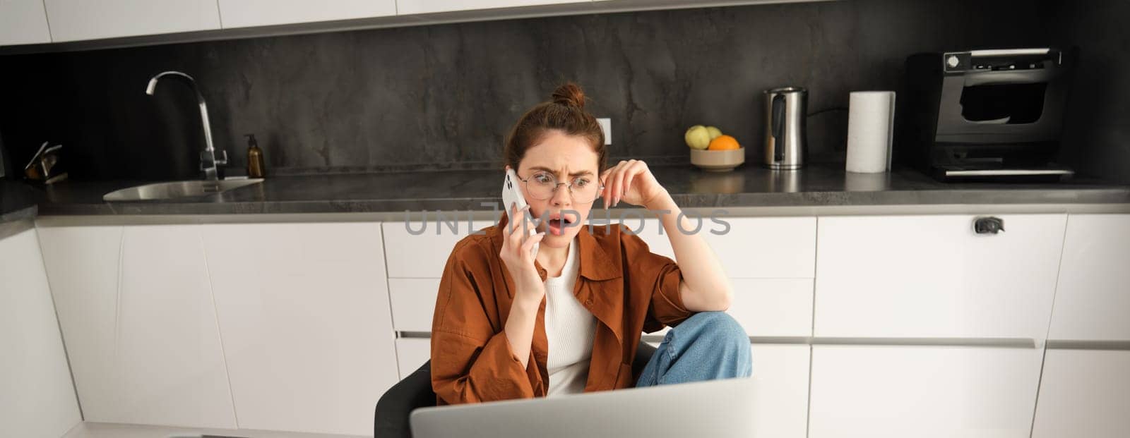 Portrait of woman with shocked face, receives concerning bad news over the phone conversation, sitting at home with laptop, working, talking to someone on telephone by Benzoix