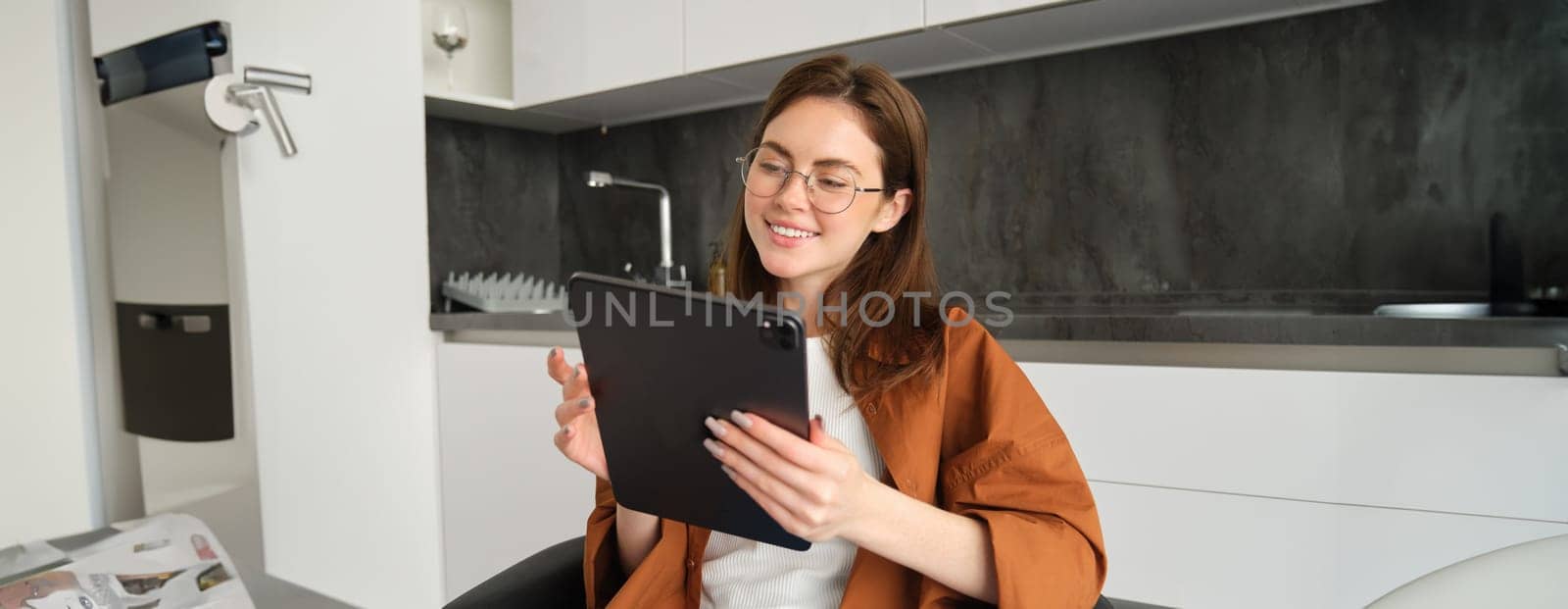 Portrait of young woman in glasses, looking at digital tablet, working from home, connects to remote class or course, watching videos online by Benzoix