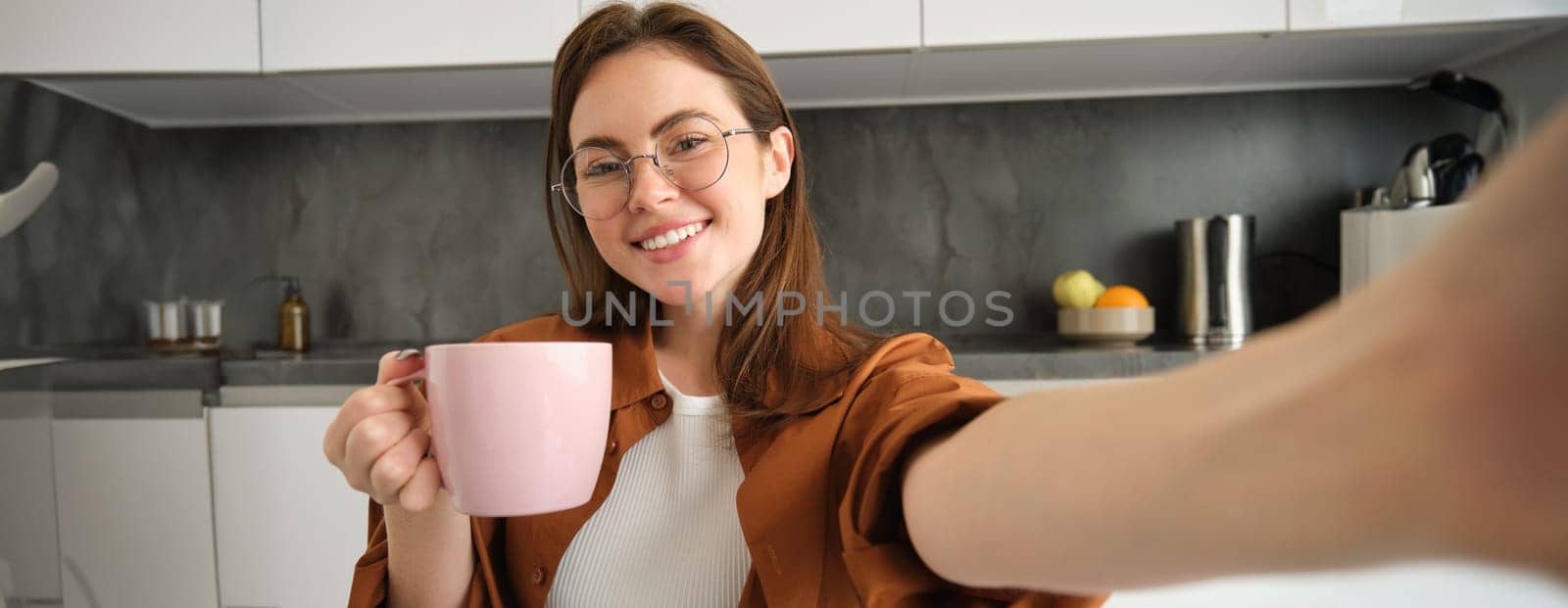 Portrait of beautiful young woman in glasses, taking selfie in kitchen with cup of morning coffee, making photos for her daily routine lifestyle blog by Benzoix