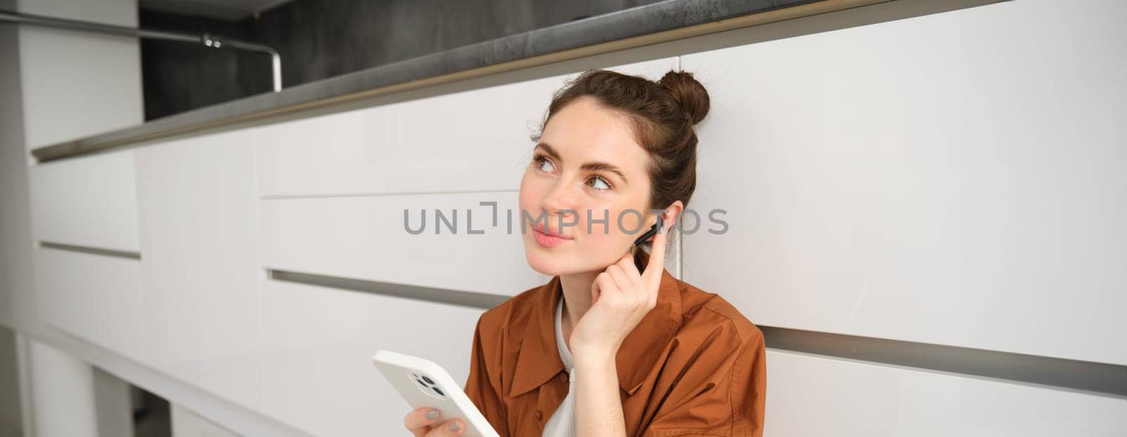 Portrait of cute young woman in wireless headphones, listening music, watching video on smartphone and using earphones, sitting on kitchen floor by Benzoix