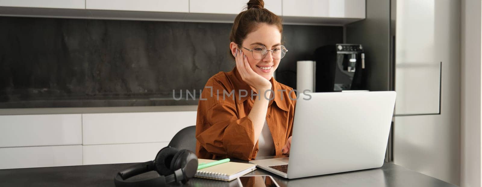 Portrait of young woman working from home, sets up workplace in kitchen, using laptop. Student doing homework on computer.