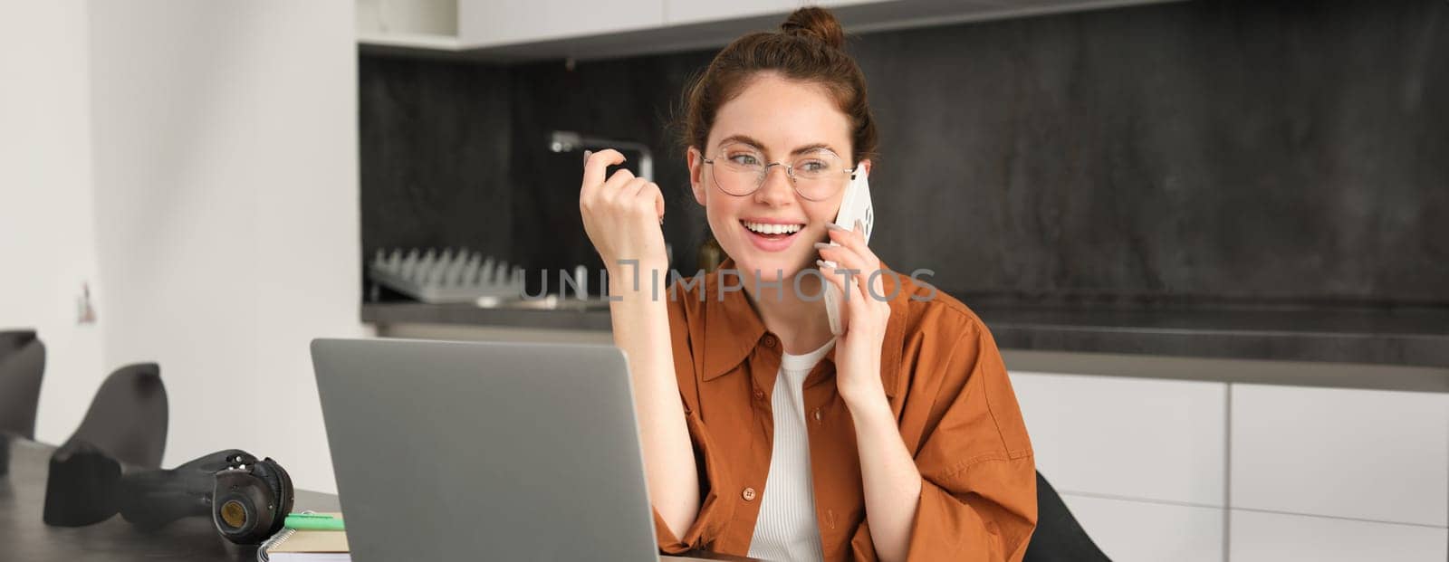 Portrait of beautiful smiling woman working from home, talking on mobile phone, calling client, self-employed businesswoman sets up workplace in kitchen, using laptop and smartphone.