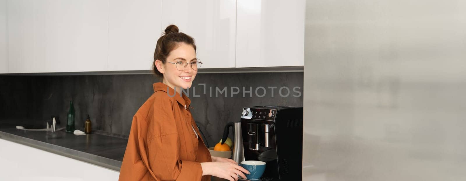 Portrait of young woman making herself coffee, standing near machine and smiling, holding mug for cappuccino, looking happy at camera.