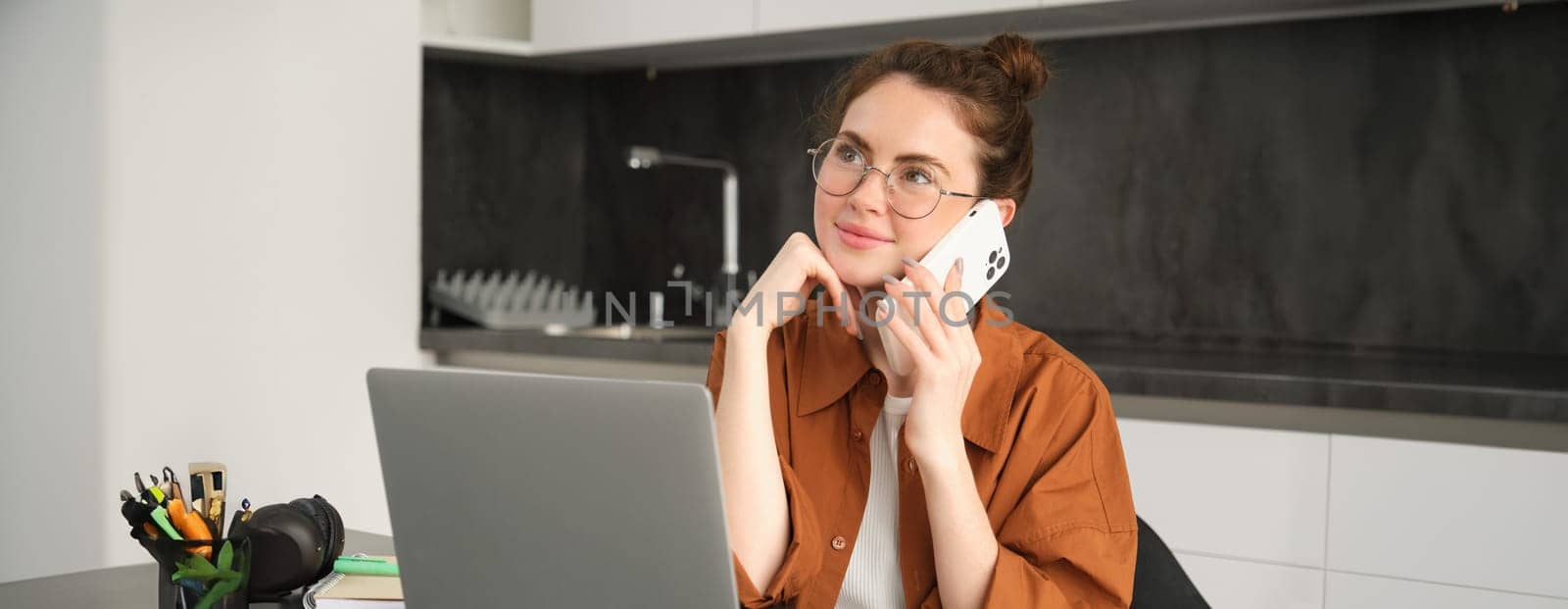 Portrait of young self-employed woman, entrepreneur working from home, freelancer calling client. Girl making an order, talking to someone on phone, sitting in kitchen with laptop by Benzoix