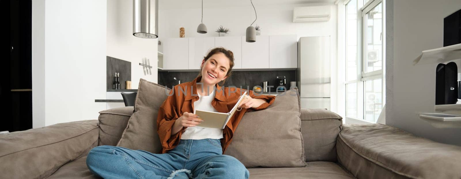 Young woman sits at home on sofa, reading her notes, holding notebook, studying for exam, student revising in her living room.