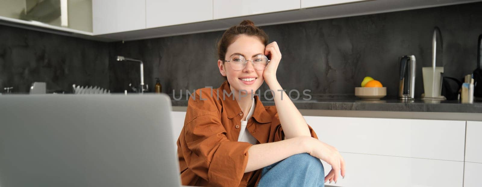 Portrait of young woman, business owner working from home, freelancer using laptop, sitting in kitchen, wearing glasses.