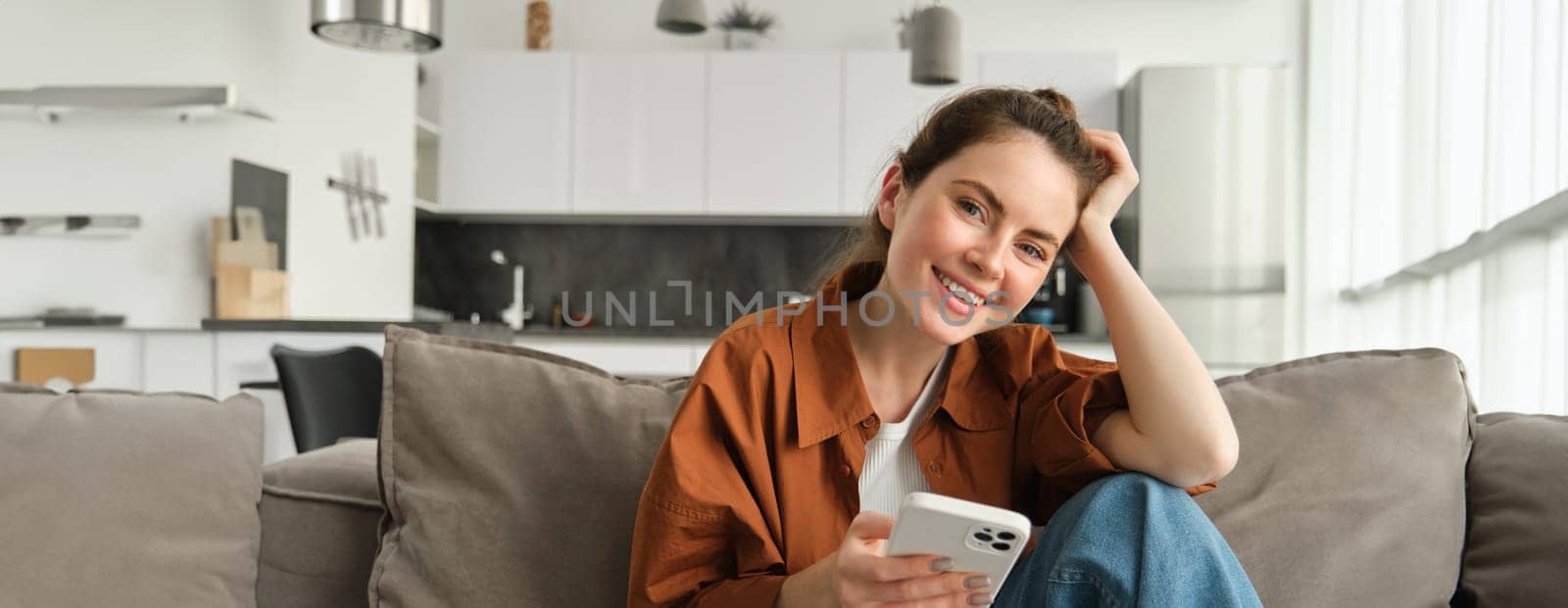 Portrait of cute young smiling woman, sitting on sofa with mobile phone, relaxing at home and using smartphone, scrolling social media, buying online, ordering something on application by Benzoix