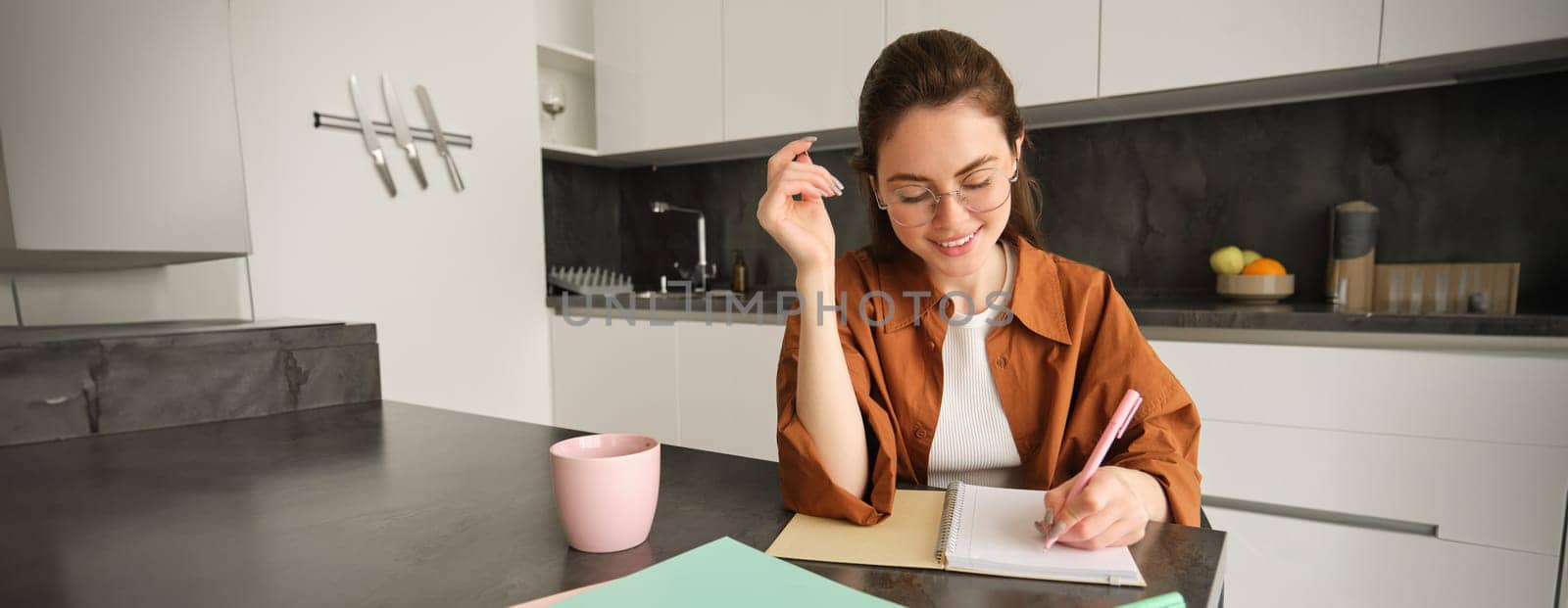 Portrait of young smiling woman, tutor preparing notes for lesson at home, writing in notebook and looking happy, student studying in kitchen.