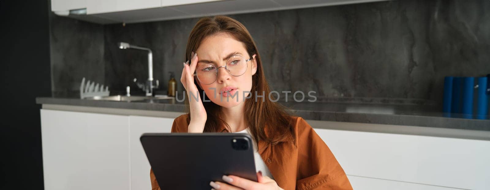 Portrait of woman freelancer, wearing glasses, holding digital tablet, looking tired, has migraine or headache, reading e-book. Freelance worker sits at home with gadget.