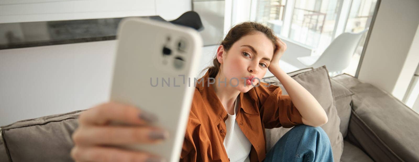 Portrait of cute young woman, modern girl taking selfies on smartphone app, posing on couch, extends her white mobile phone to take picture, pucker lips for photo by Benzoix