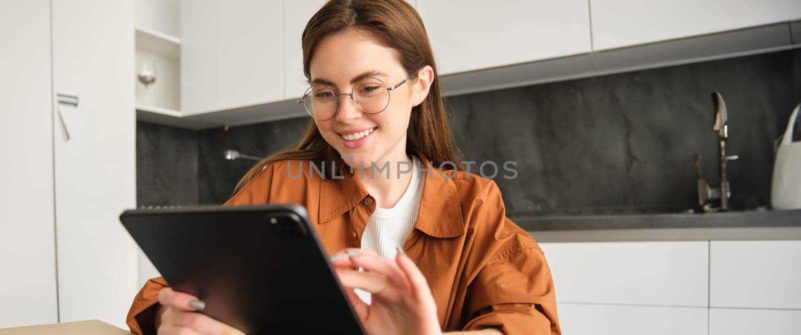Portrait of young woman in glasses, beautiful girl student reading on digital tablet, messaging, doing online shopping while sitting in kitchen.