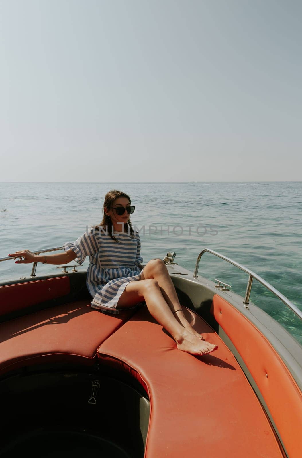Portrait of one beautiful Caucasian brunette girl in a striped dress and sunglasses sitting on the stern of a boat while sailing against the backdrop of a blurry blue sea on a sunny summer day, close-up side view.