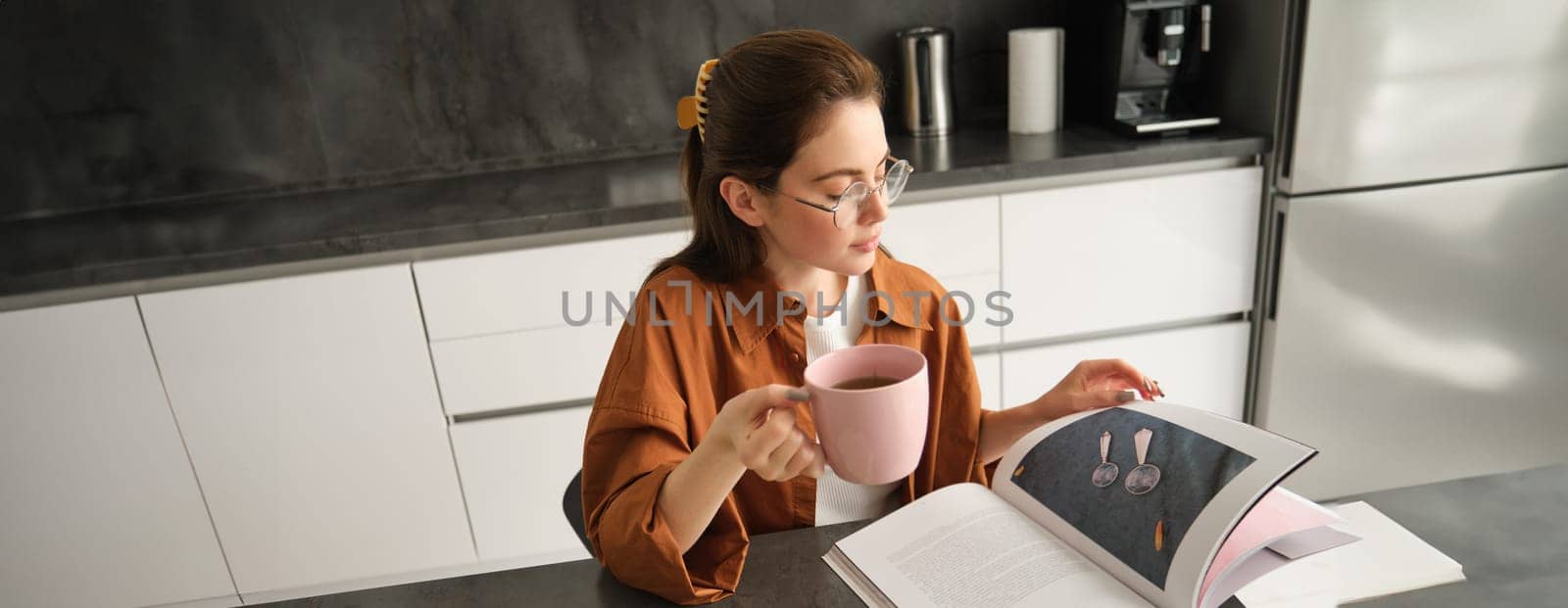 Portrait of beautiful young woman in kitchen, sitting with a book, flipping pages, reading and drinking coffee.