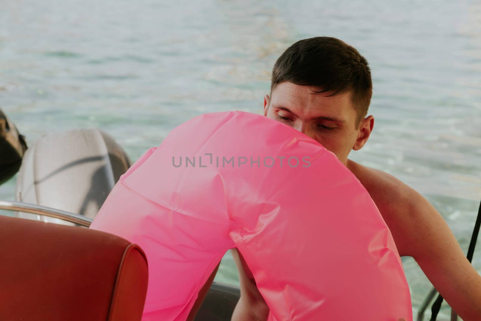 Portrait of one young handsome Caucasian brunette inflates a pink lifebuoy with his mouth while sitting in a boat against the backdrop of a blurred sea on a sunny summer day, close-up side view.