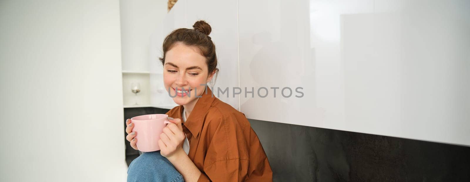 Portrait of young woman with cup of coffee, sits in kitchen and drinks aromatic drink at home, holds tea mug by Benzoix