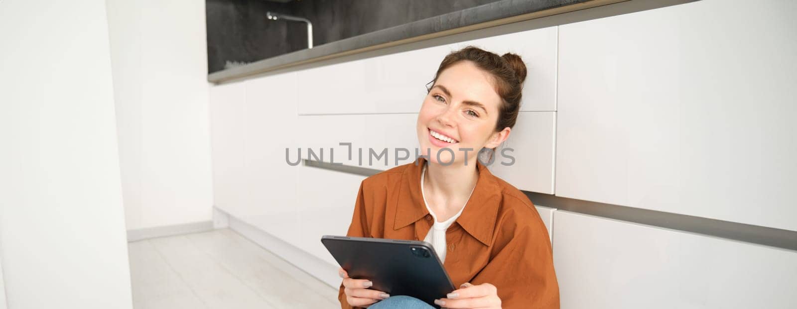 Portrait of young beautiful woman sitting on kitchen floor with digital tablet, browsing news feed, social media app on gadget, smiling and looking happy by Benzoix