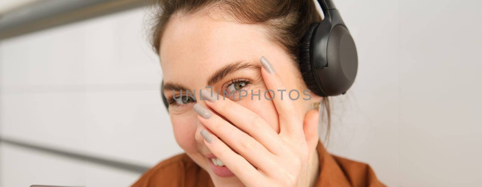 Cute smiling girl, blushes, hides face behind hand and laughing, wears wireless headphones.