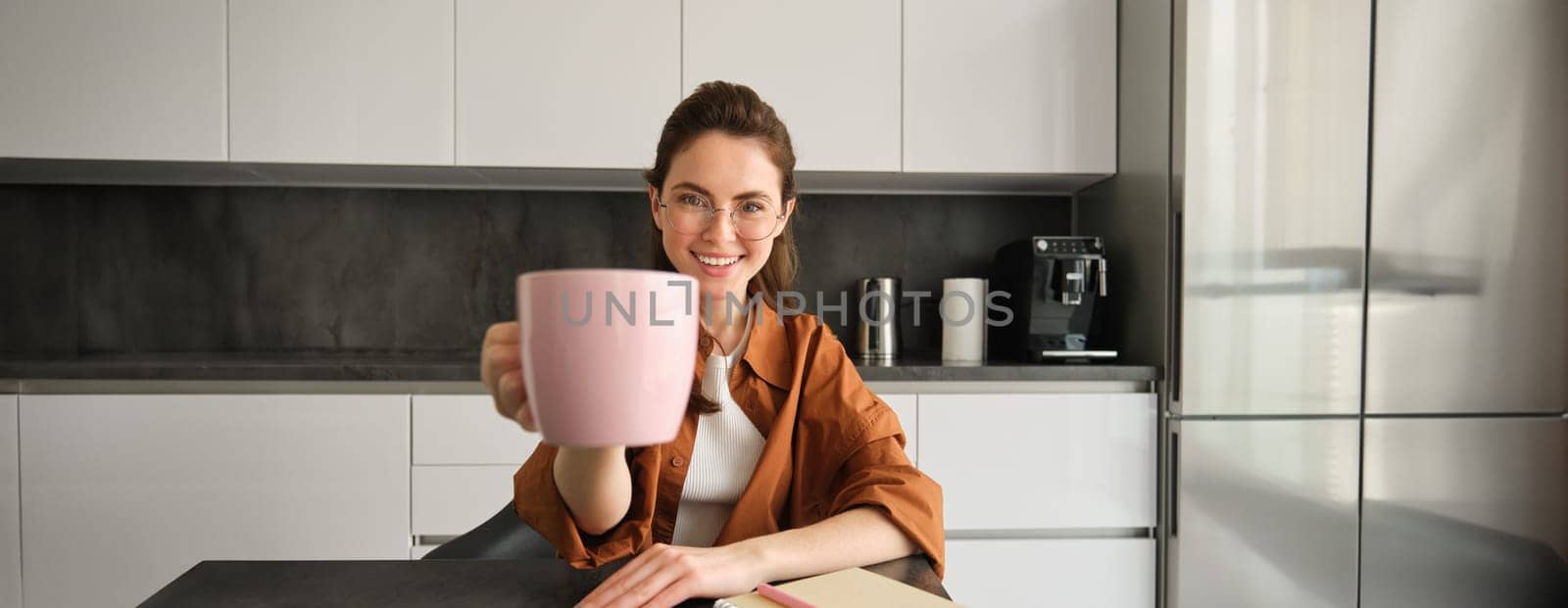 Stylish young woman, student studying from home, sitting in kitchen with homework, giving you cup of coffee, raising pink mug, smiling at camera by Benzoix