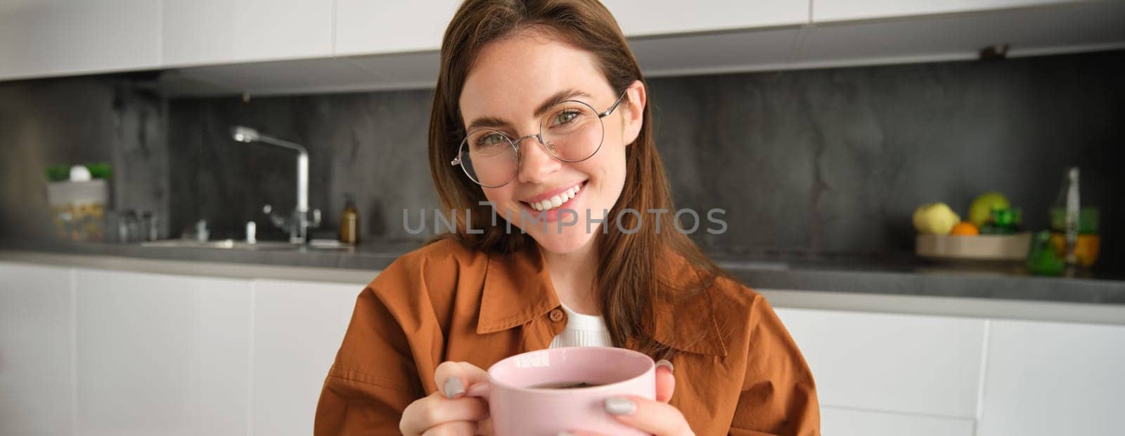 Close up portrait of beautiful girl in glasses, holding hot cup of drink, drinking tea or coffee at home and smiling at camera.