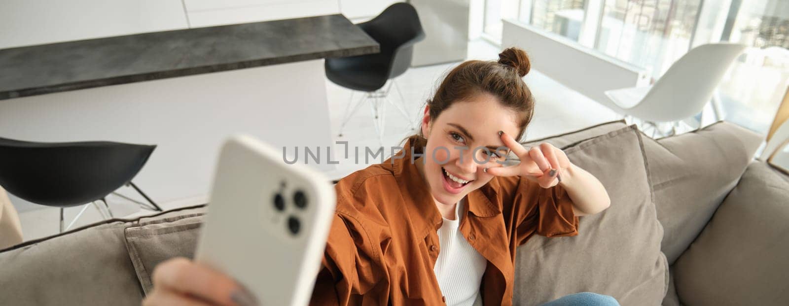 Positive and happy young woman taking selfie on smartphone, sitting on couch, showing peace, v-sign gesture by Benzoix