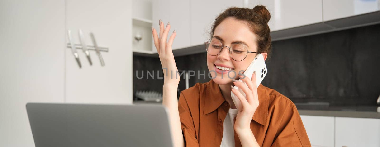 Portrait of young businesswoman, self-employed lady working from home, making phone calls to clients and smiling, using laptop in kitchen.
